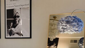 A room with a brown sofa, a white armchair, a large mirror reflecting a chandelier, and a framed newspaper on the wall. - 3 KING Beds & Free parking • Central Place (Pittsburgh)