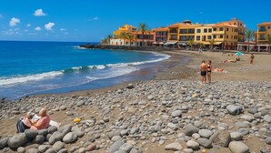 Une plage au rivage rocheux, un ciel bleu dégagé et des bâtiments visibles à l’arrière-plan.