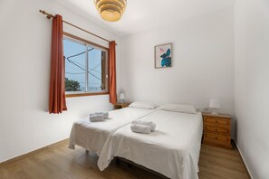 A bedroom with two beds, a window with curtains, a bedside table with a lamp, and a wall-mounted picture.