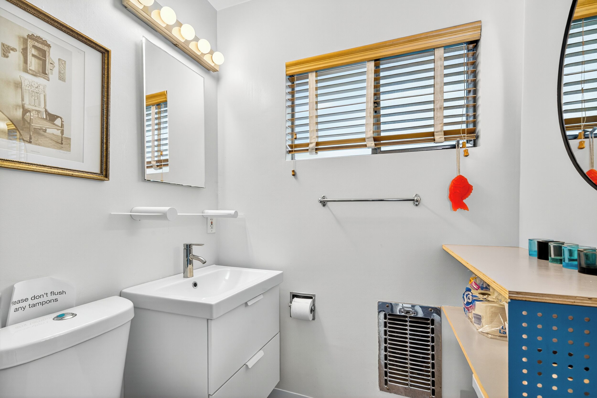 Un baño moderno con lavamanos, inodoro y tocador. Cuenta con una ventana con persianas, un espejo con luces y un cuadro enmarcado en la pared.
