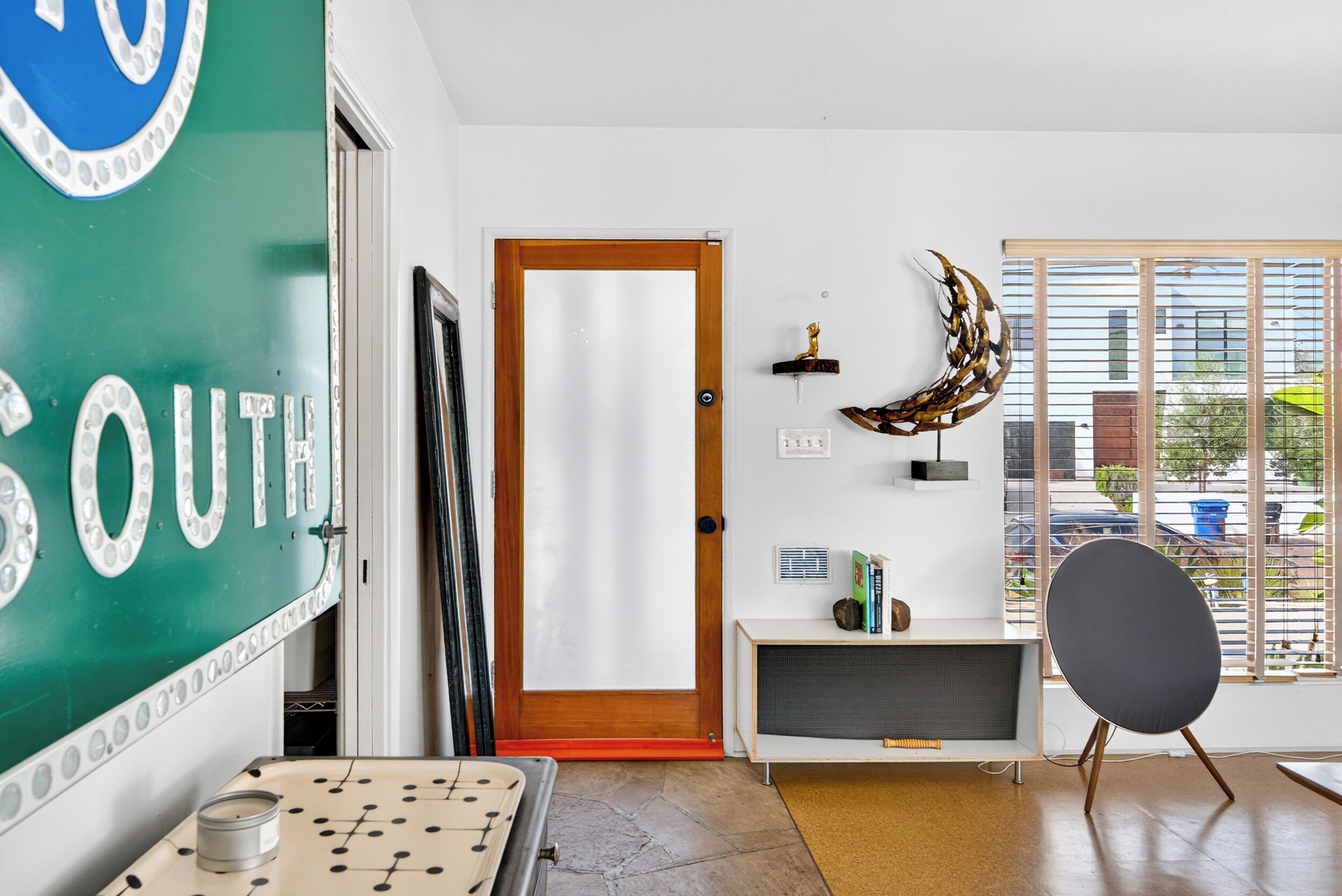 A room with a green wall, a wooden door, a black chair, a small shelf with books, and a window with blinds.