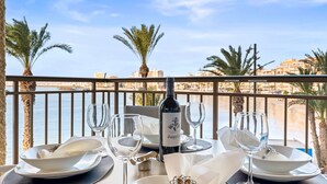 A table set for dining with a bottle of wine, empty glasses, and plates, overlooking a beach with palm trees and buildings in the background.
