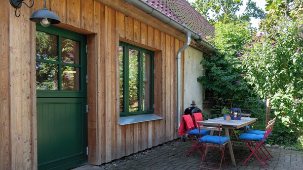 Un edificio in legno con una porta e finestre verdi, un patio in mattoni e un tavolo con sedie blu.