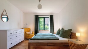 Una camera da letto con un letto di legno, una cassettiera bianca, una poltrona gialla, una scrivania con una lampada e una finestra con tende.
