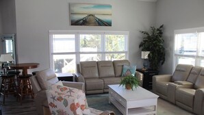 A living room with a beige sofa, a white coffee table, and a wicker chair. There is a painting on the wall and a plant on the coffee table. - ISLAND PARK - SECOND ROW! AVAILABLE NOW! By Williamson Realty (Ocean Isle Beach)