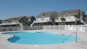 A swimming pool with a wooden deck, surrounded by a white fence, and adjacent to a row of beach houses with balconies. - ISLAND PARK - SECOND ROW! AVAILABLE NOW! By Williamson Realty (Ocean Isle Beach)