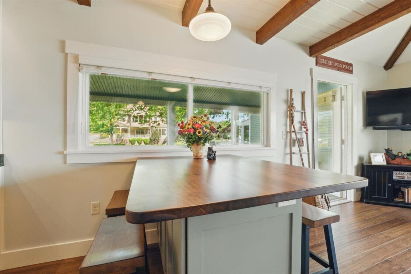 Una cocina con una isla de madera, un ventanal con vistas a la vegetación y un cartel que dice "COME SIT ON MY PORCH".