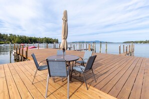 Une terrasse en bois avec une table ronde et des chaises, un parasol, et une vue sur l’eau et les bateaux.