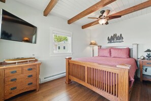 Une chambre à coucher avec un lit en bois, une table de chevet, une commode, un ventilateur de plafond, une fenêtre donnant sur une maison et un téléviseur fixé au mur.