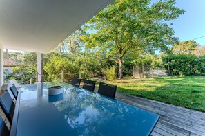 Een eethoek buiten met een glazen tafel en stoelen, omgeven door een houten terras en een gazon met bomen.