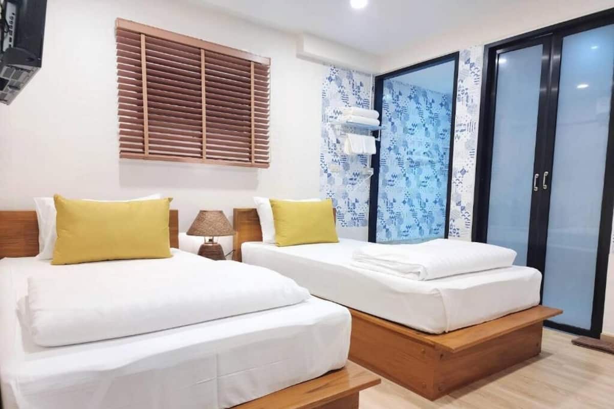 A hotel room with two beds, wooden headboards, a window with blinds, a wall-mounted air conditioning unit, and a sliding glass door.