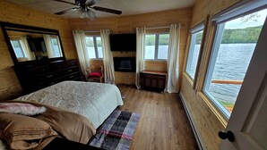 Un dormitorio de cabaña de madera con una cama, una silla, un televisor y una ventana con vista a un lago.