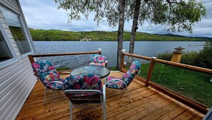 Una terraza de madera con una mesa y sillas, con vista a un lago y colinas boscosas.