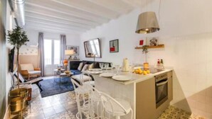 A modern living room with a sofa, dining table, and chairs. There is a kitchen area with a sink and a stove. The room has a large window and a ceiling with exposed beams. - Boutique Living in the Heart of Barcelona (Barcelona)