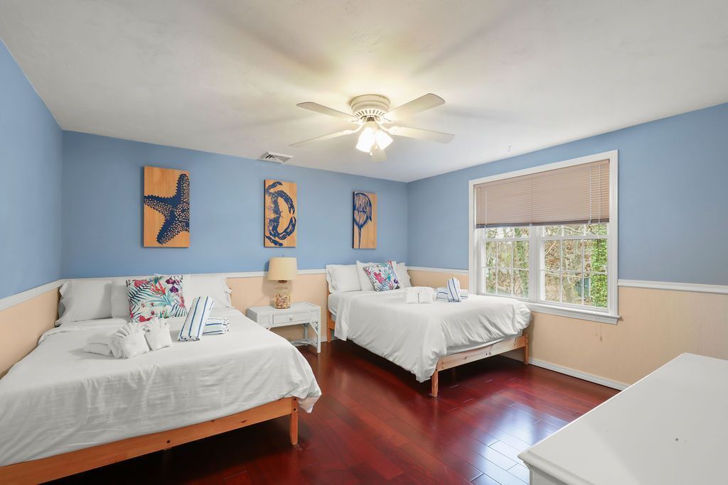 A bedroom with two beds, a ceiling fan, artwork on the walls, a window with blinds, and a bedside table with a lamp.