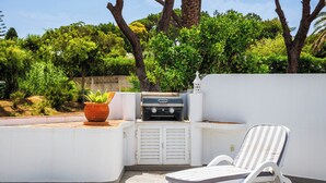 Une terrasse extérieure ensoleillée, équipée d’un barbecue, d’un fauteuil de relaxation et d’une plante en pot.
