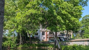 A wooden dock extending over a calm body of water, surrounded by lush greenery and a multi-story building in the background.