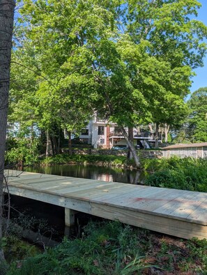Un muelle de madera se extiende sobre una masa de agua tranquila, rodeado de exuberante vegetación y un edificio de varios pisos al fondo.