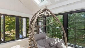 A large room with a hanging chair, large windows, and a view of greenery outside.