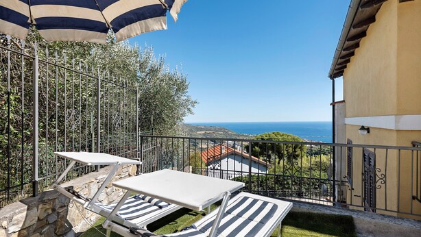 Een balkon met een tafel en twee gestreepte lounge fauteuils, een gestreepte parasol, en uitzicht op zee en verre gebouwen.