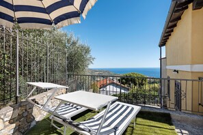 Ein Balkon mit Tisch und zwei gestreiften Liegestühlen, einem gestreiften Sonnenschirm und Blick auf das Meer sowie entfernte Gebäude.