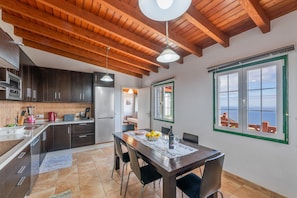 A kitchen with wooden beams, a dining table with chairs, a refrigerator, and a view through a window.