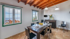 Un comedor con techo de madera, ventanas amplias, una cocina con televisor y una mesa dispuesta con un bol de fruta.