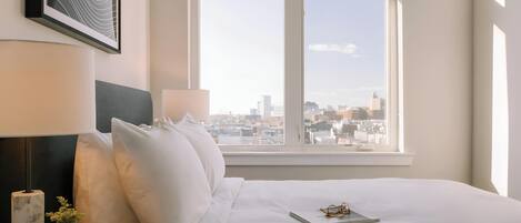 Une chambre d’hôtel avec une grande fenêtre, un lit avec du linge de lit blanc, une table de chevet avec une lampe, un vase de fleurs et un tableau encadré au mur.