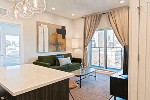 A modern living room with a sofa, a coffee table, and a TV. There is a large window with curtains and a city view.