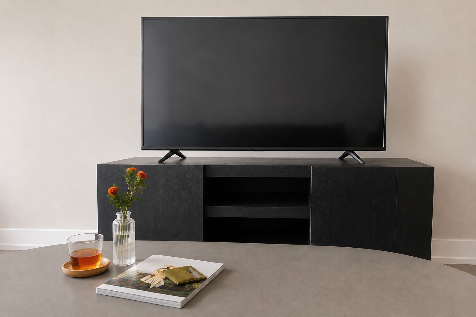 A modern living room with a flat-screen TV mounted on a dark wooden cabinet, a glass vase with flowers, a book, and a glass of tea on a round table.