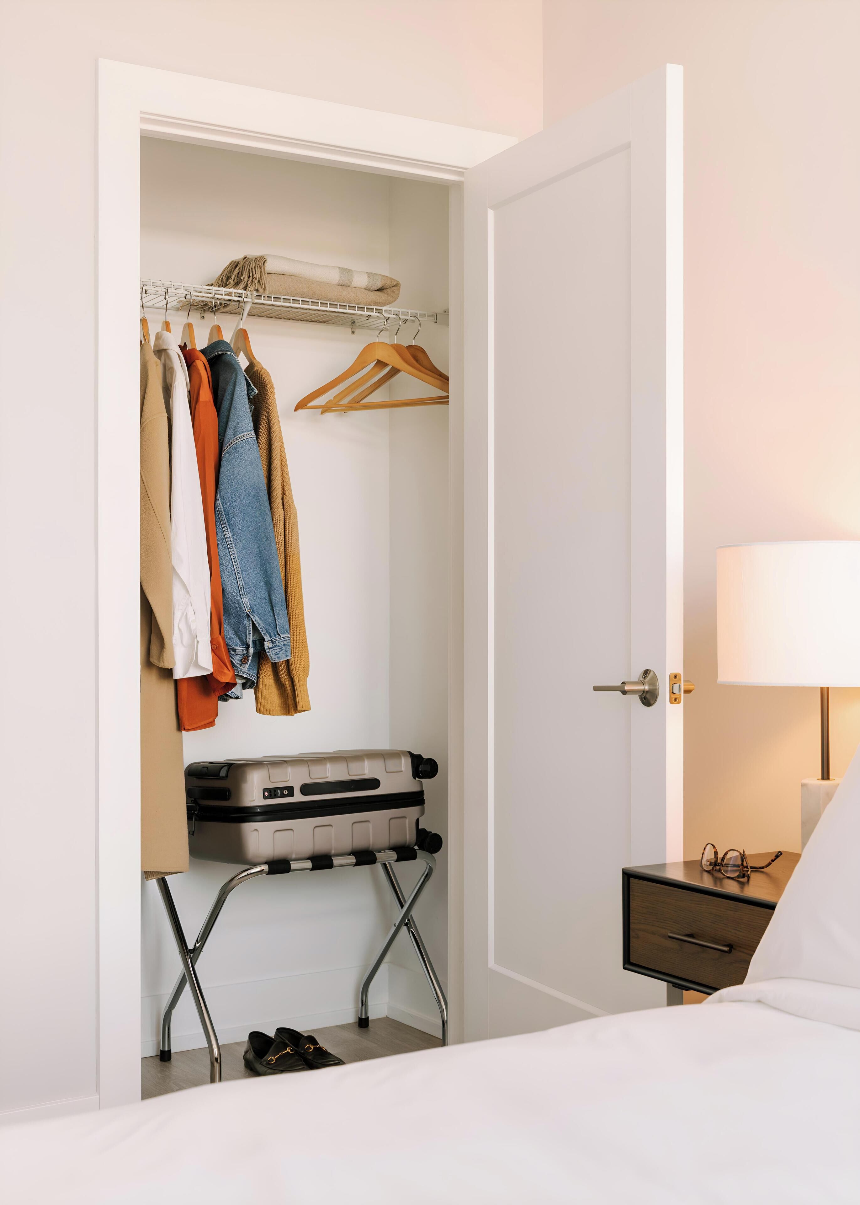 A hotel room with a wardrobe containing clothes, a suitcase on a trolley, a bedside table with a lamp, and a bed.