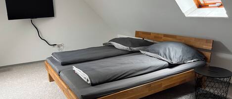 Una cama individual con estructura de madera y ropa de cama gris, una mesita redonda con base de alambre negra, y un televisor montado en la pared.