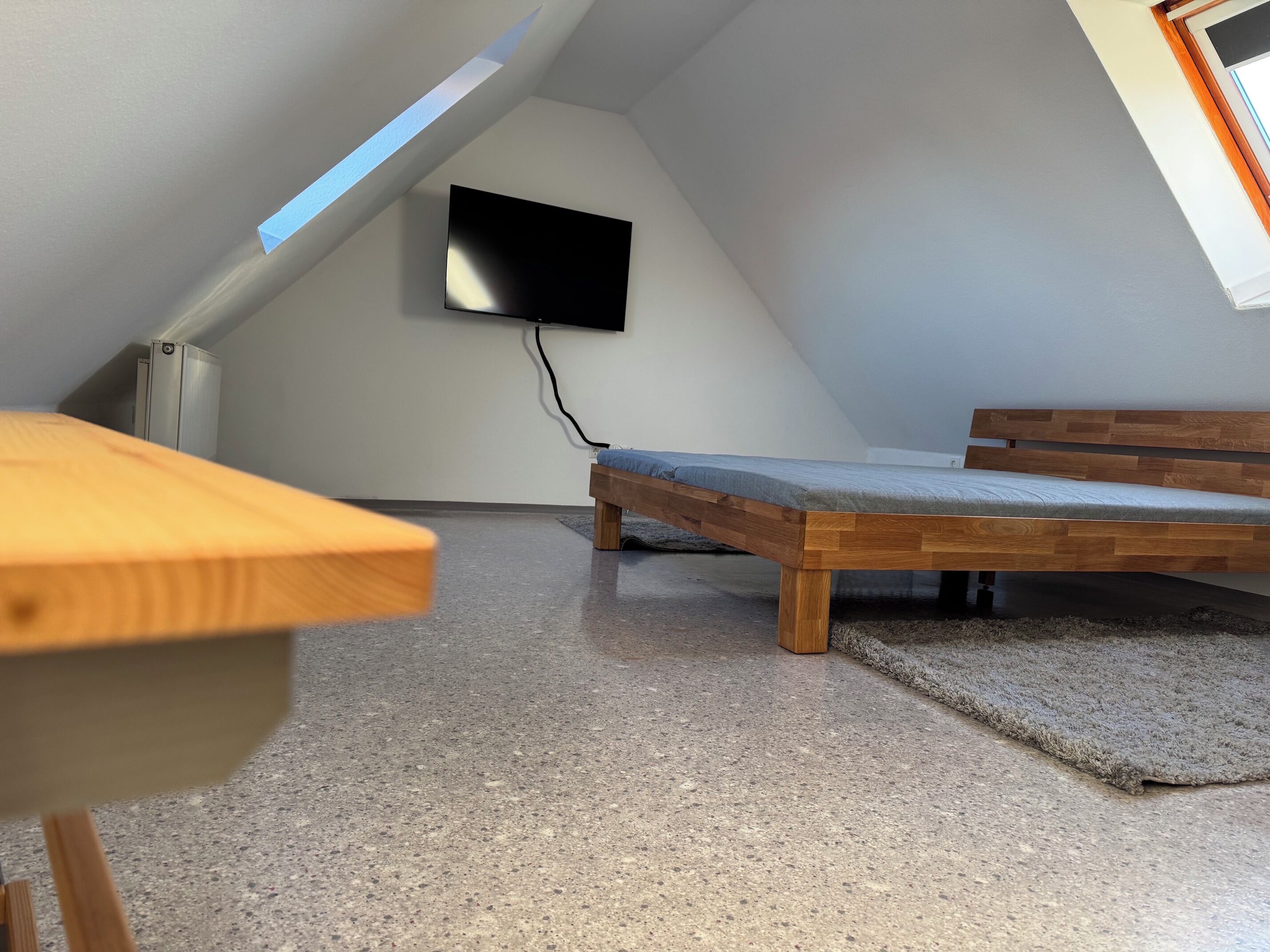 Een kamer met een laag schuin dak, een bed, een tafel en een aan de muur bevestigde televisie.