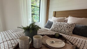 Ein ordentlich gemachtes Bett mit karierter Bettdecke, ein Tablett mit zwei Tassen und einem Teller, sowie ein Fenster mit Vorhängen.
