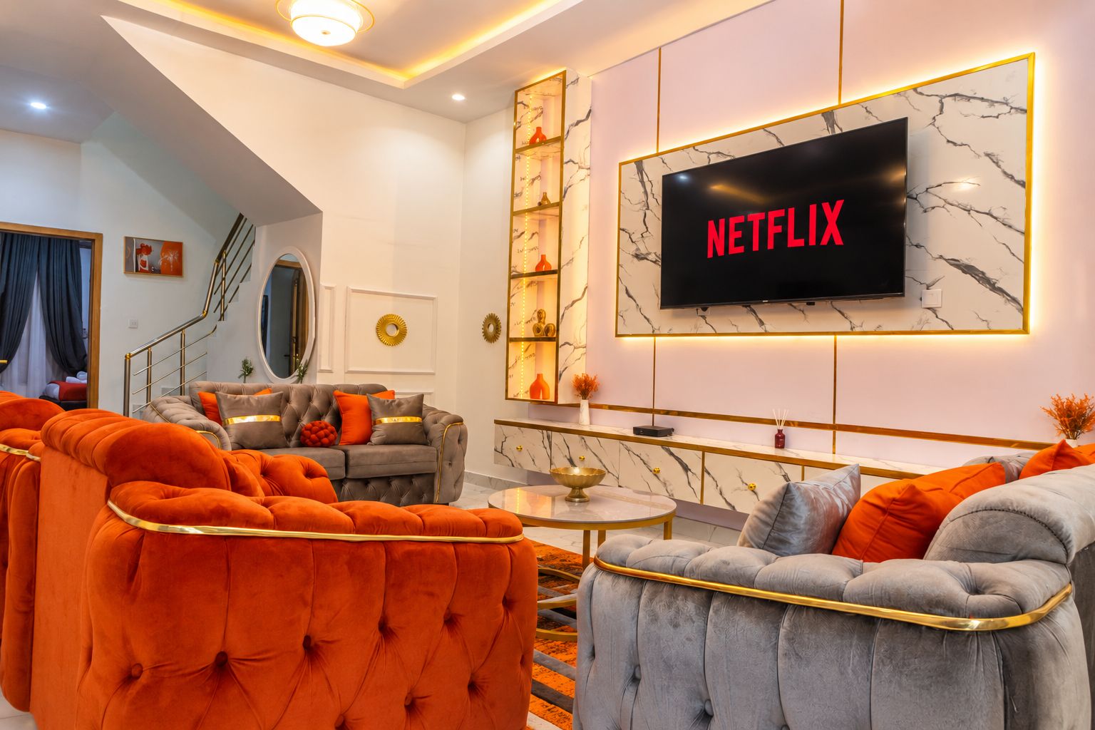 A modern living room with a large flat-screen TV mounted on the wall, a grey sofa with orange cushions, and a round coffee table.