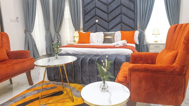 A modern hotel room with a large bed, two orange armchairs, a round coffee table, and a grey upholstered headboard. - Fortified 4 Bedroom Home (Lekki)