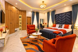 A modern hotel room with a large bed, a sitting area with a sofa and armchairs, a desk, and a television. - Fortified 4 Bedroom Home (Lekki)