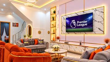 Una sala de estar moderna con un televisor grande mostrando el logo de la Premier League, un sofá gris, sillones naranja y una pared con estampado de mármol.