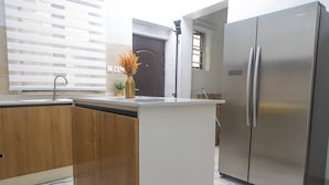 A modern kitchen with a central island, stainless steel refrigerator, wooden cabinets, and a window with blinds. - Fortified 4 Bedroom Home (Lekki)