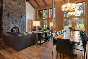 A spacious room with a stone fireplace, a dining table with chairs, and large windows offering a view of the outdoors.