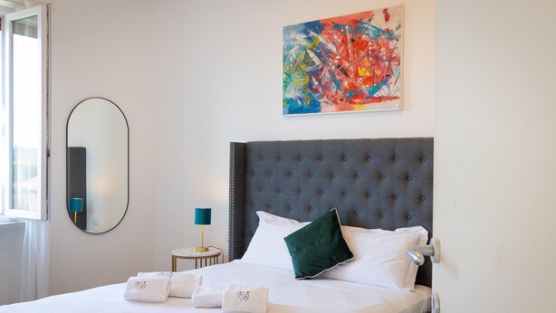 A neatly made bed with white linens and pillows, a large mirror, a colorful abstract painting, and a small bedside table with a lamp. - Rental in Rome Vatican Secret Apartment (Rome)