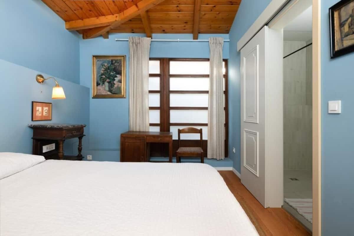 A bedroom with a bed, a desk, a chair, a painting, and a window with curtains.