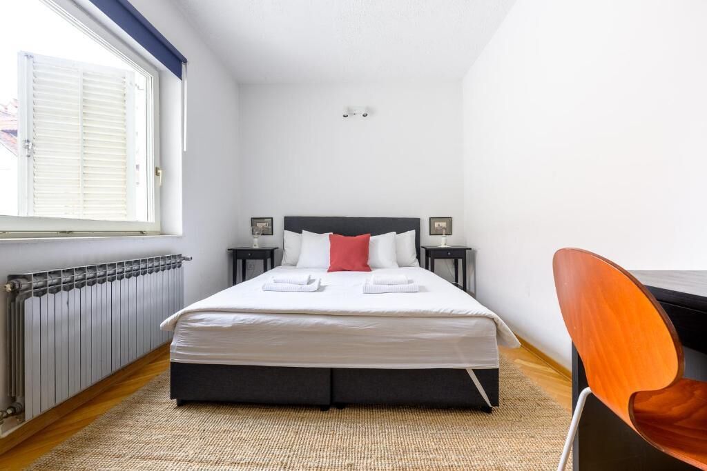 A bedroom with a bed, two bedside tables, a window with blinds, a radiator, and a chair.