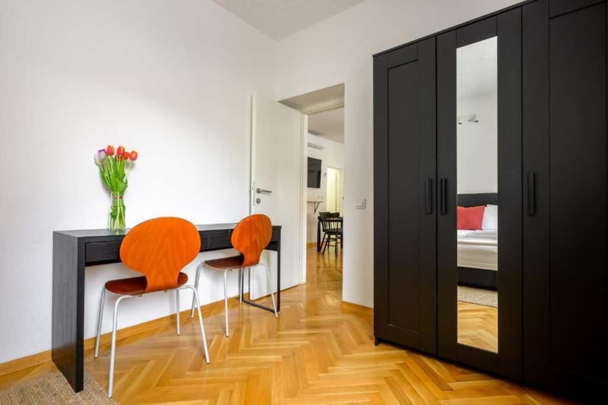 A modern room with a wooden herringbone floor, a black wardrobe with a mirror, a desk with a chair, and a vase of tulips.