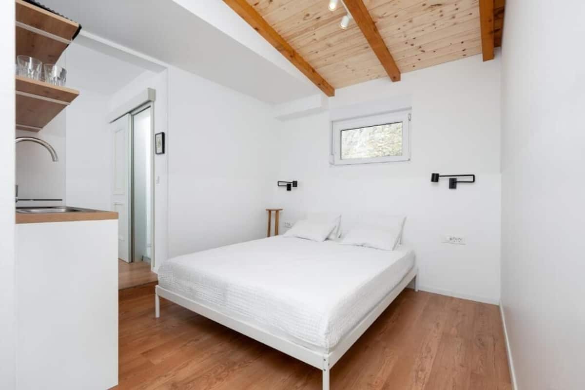 A bedroom with a bed, wooden beams, a window, and a kitchen area.