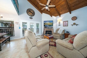 A living room with a fireplace, two armchairs, a sofa, a wooden ceiling fan, and a dining area with a bench and table.