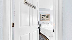 Una puerta blanca con un letrero que dice “THE SEAGLASS SHACK” da acceso a un dormitorio con una cama, un cuadro enmarcado en la pared y piso de madera.