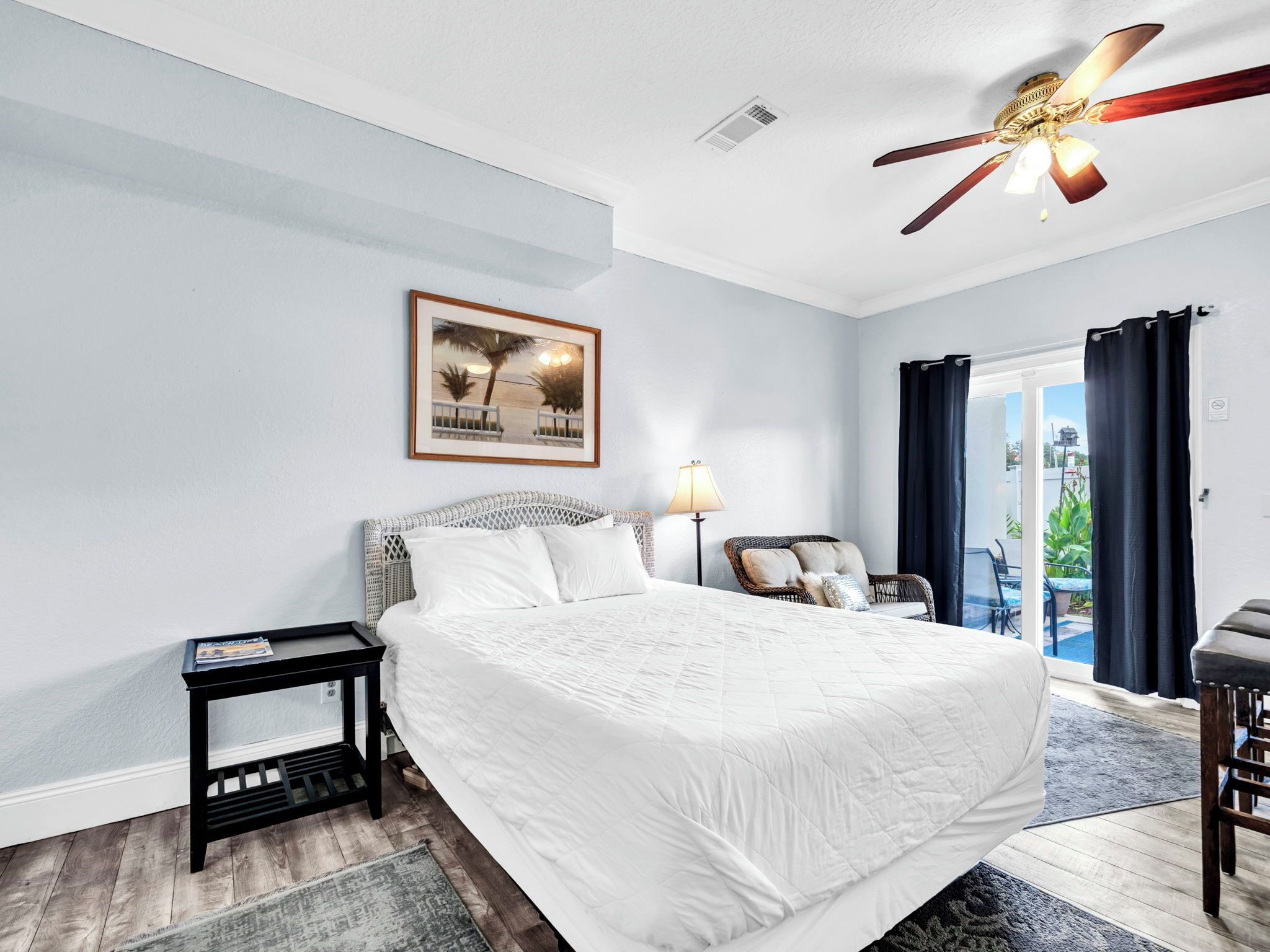 A bedroom with a large bed, a nightstand, a lamp, a ceiling fan, and a view of the outdoors.