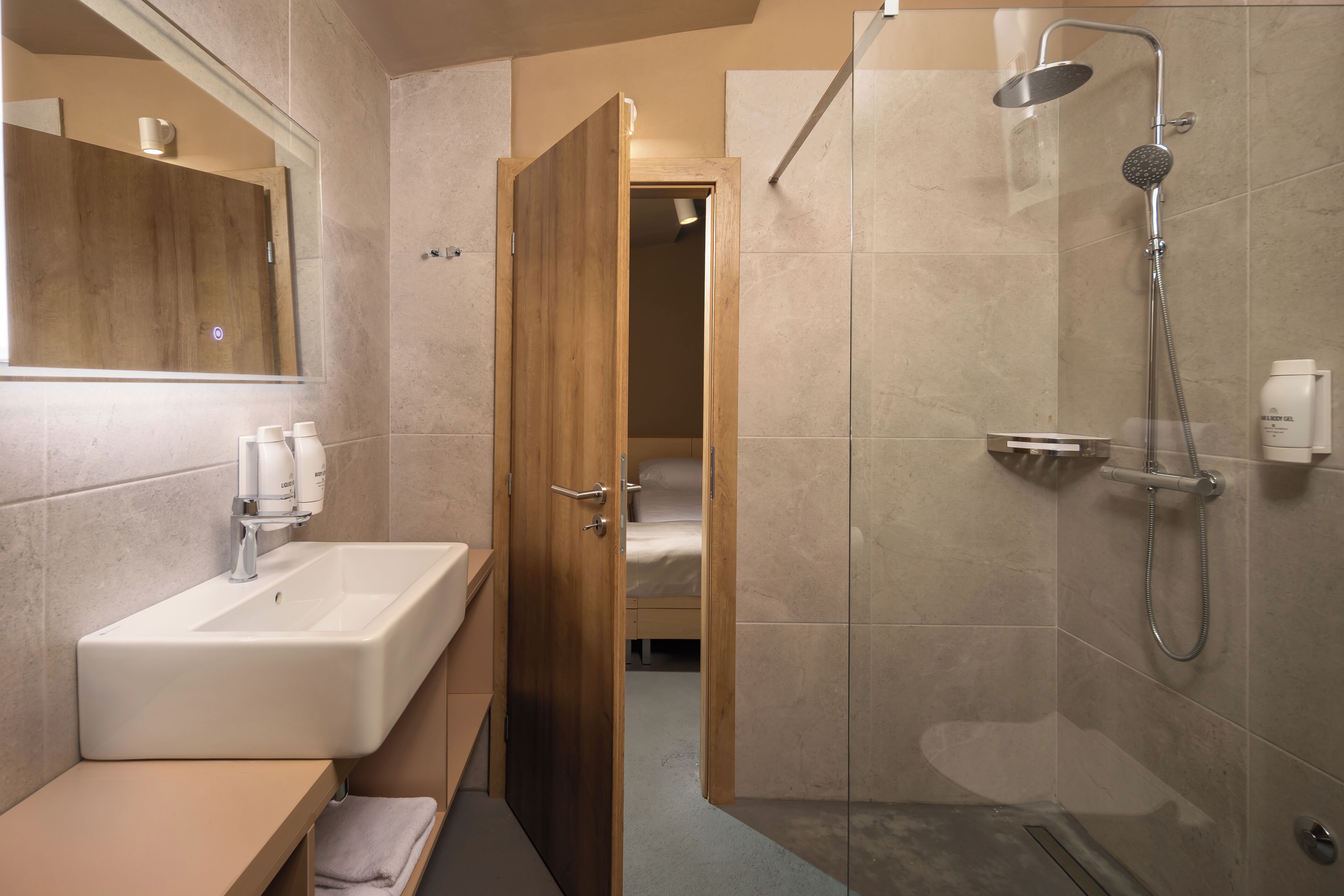Een moderne badkamer met een wastafel, spiegel, douche en een houten deur die naar een slaapkamer leidt.