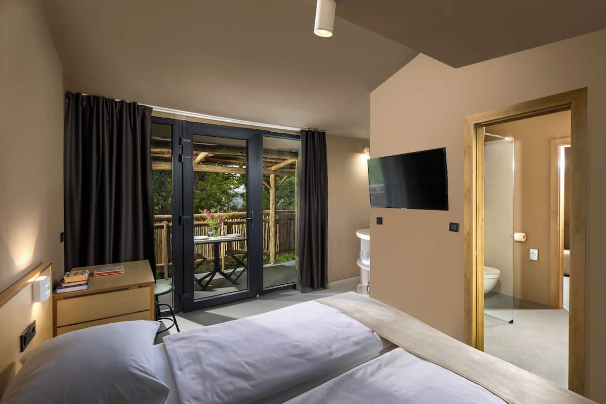 A hotel room with two beds, a desk, a TV, and a sliding glass door leading to an outdoor area with a table and chairs.